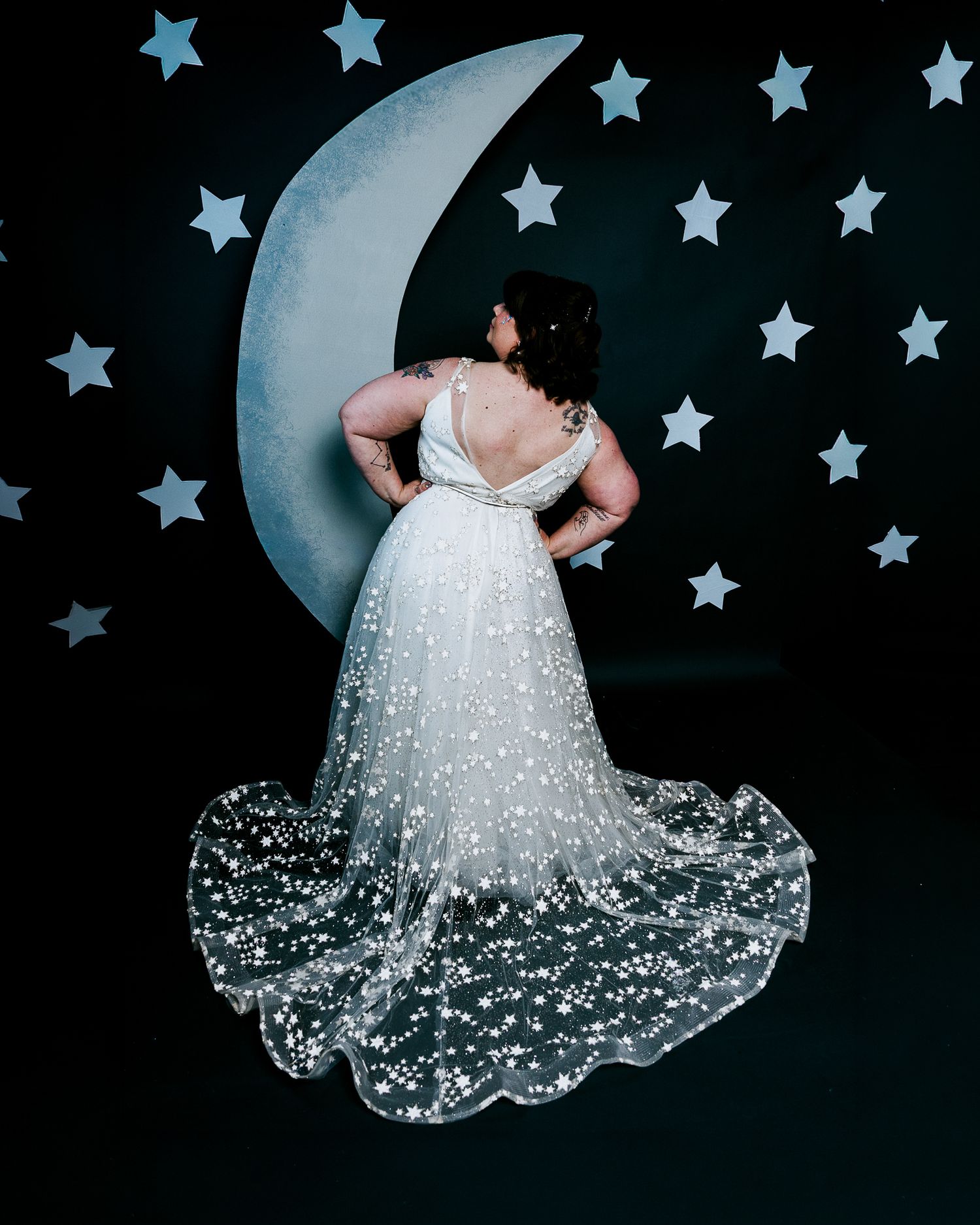 Woman in star-studded white wedding gown poses before crescent moon backdrop with star decorations.