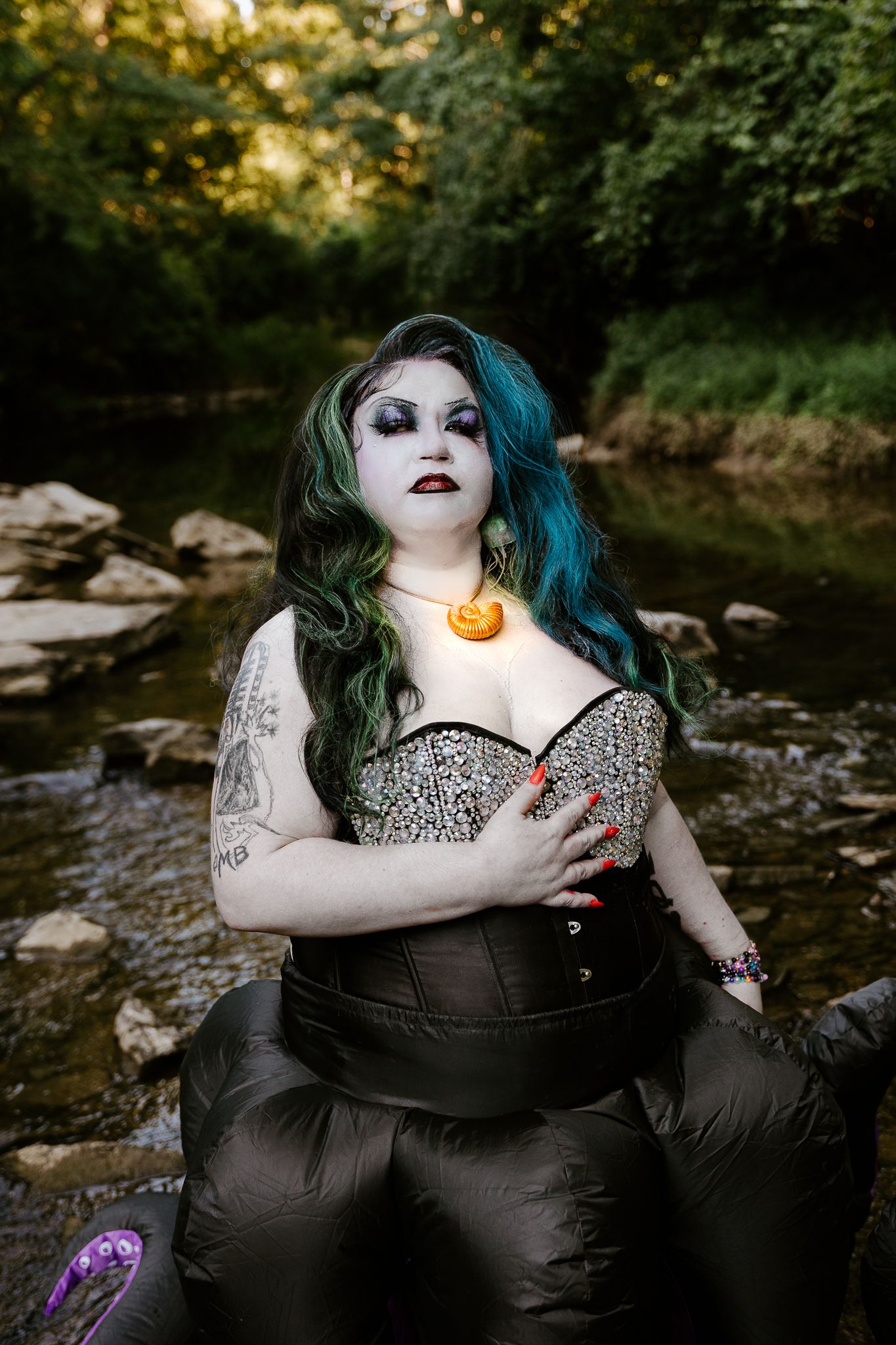 Goth woman with teal hair, dramatic makeup, corset top, glowing necklace, tattoos, posing by a rocky creek in a forest.