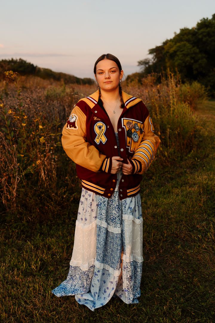 A figure in a varsity jacket and blue floral dress poses in a field at golden hour.