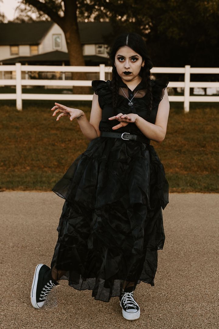 A person in a black ruffled dress and Converse sneakers poses on a path in front of a white fence.