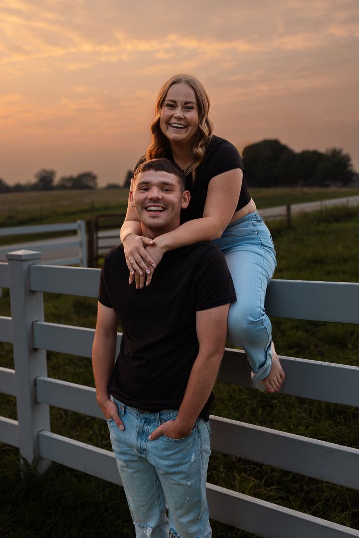 A playful couple laughs and poses together along a white fence during a golden sunset.