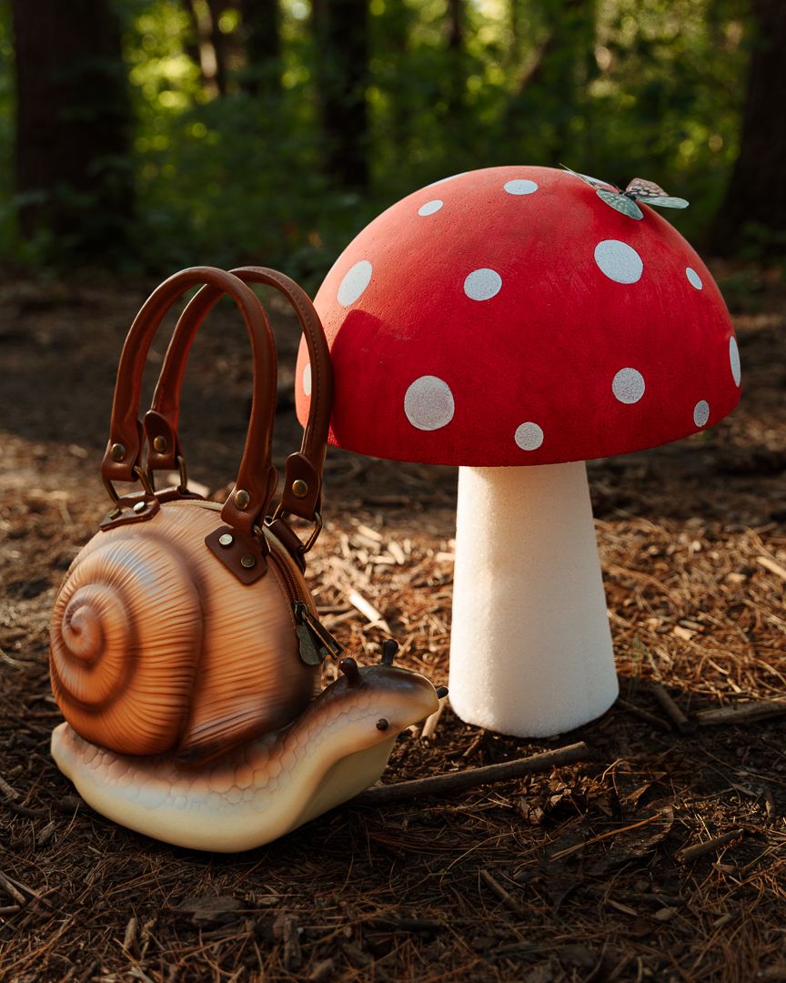 A realistic snail-shaped handbag with brown leather straps sits beside a red polka-dot mushroom sculpture in a forest.