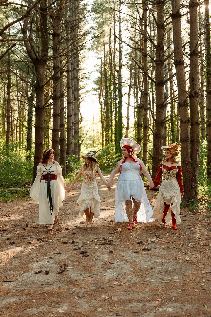 Four women in fantasy costumes walk through a sunlit pine forest, wearing white and red medieval-style dresses.