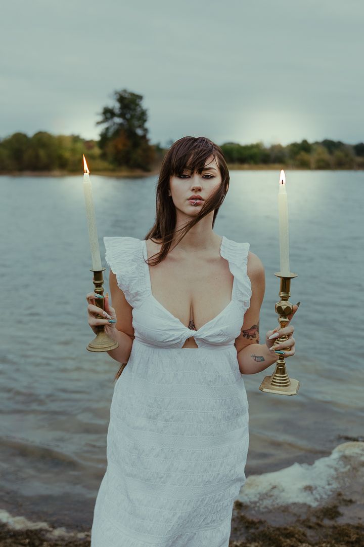 A figure in a white dress holds a lit candle by a serene lakeside at dusk.