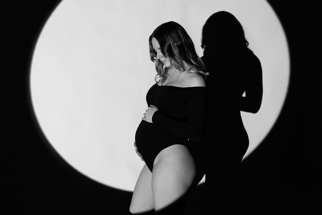 Artistic black and white silhouette of an expectant mother posing against a dramatic circular light backdrop.