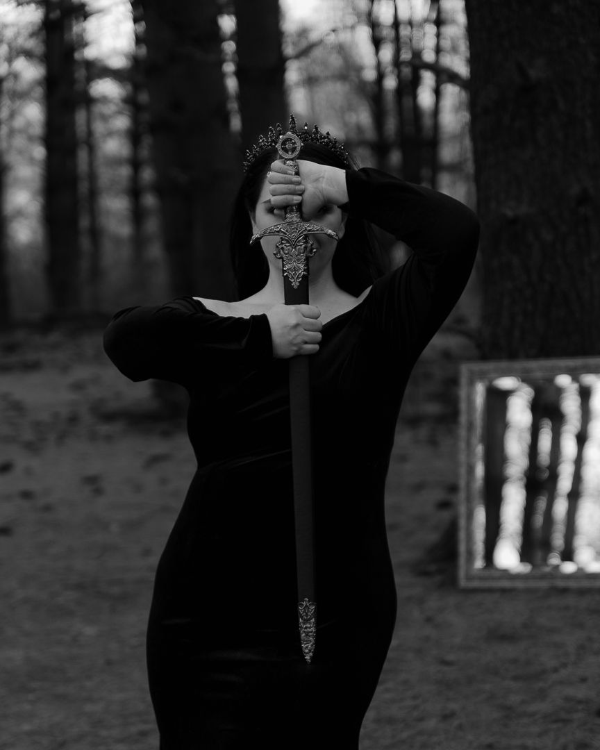 Artistic black and white photo of a figure in black holding a decorative sword in front of their face in a dark forest.
