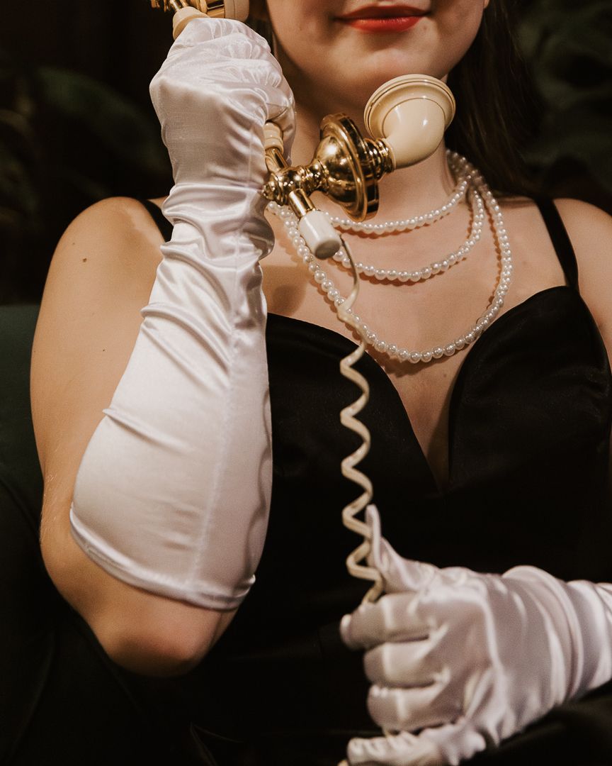 A close-up shot of white satin gloves and pearl necklace details in vintage glamour style.