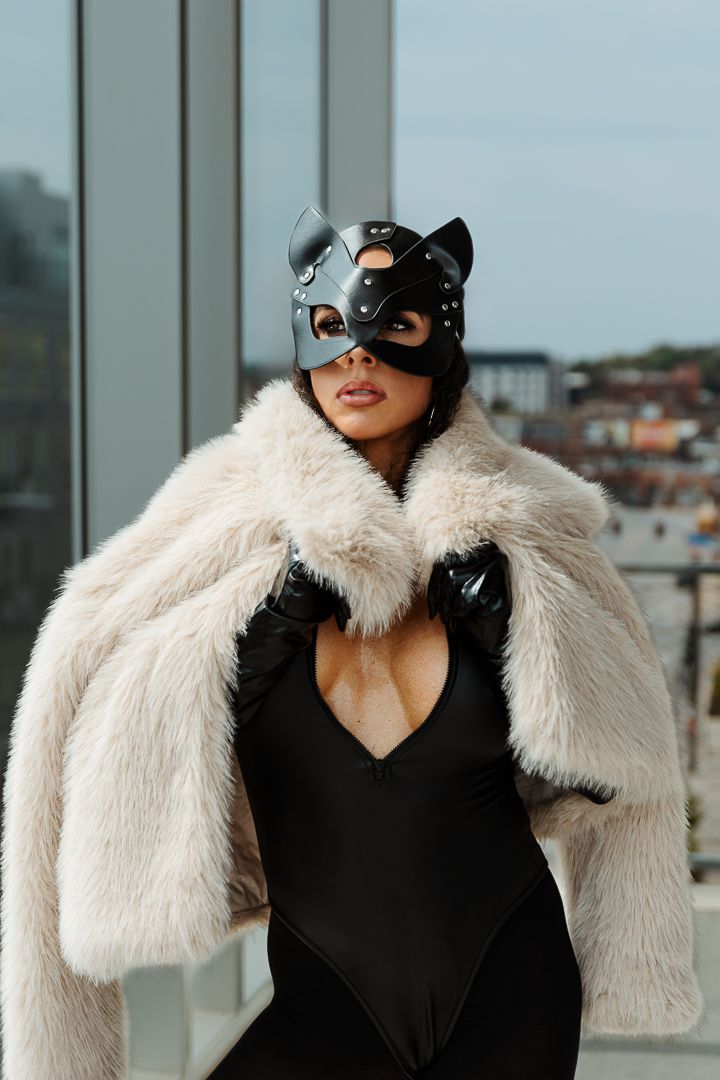 Costumed figure in black bodysuit and white fur coat poses dramatically on urban rooftop wearing cat mask and gloves.