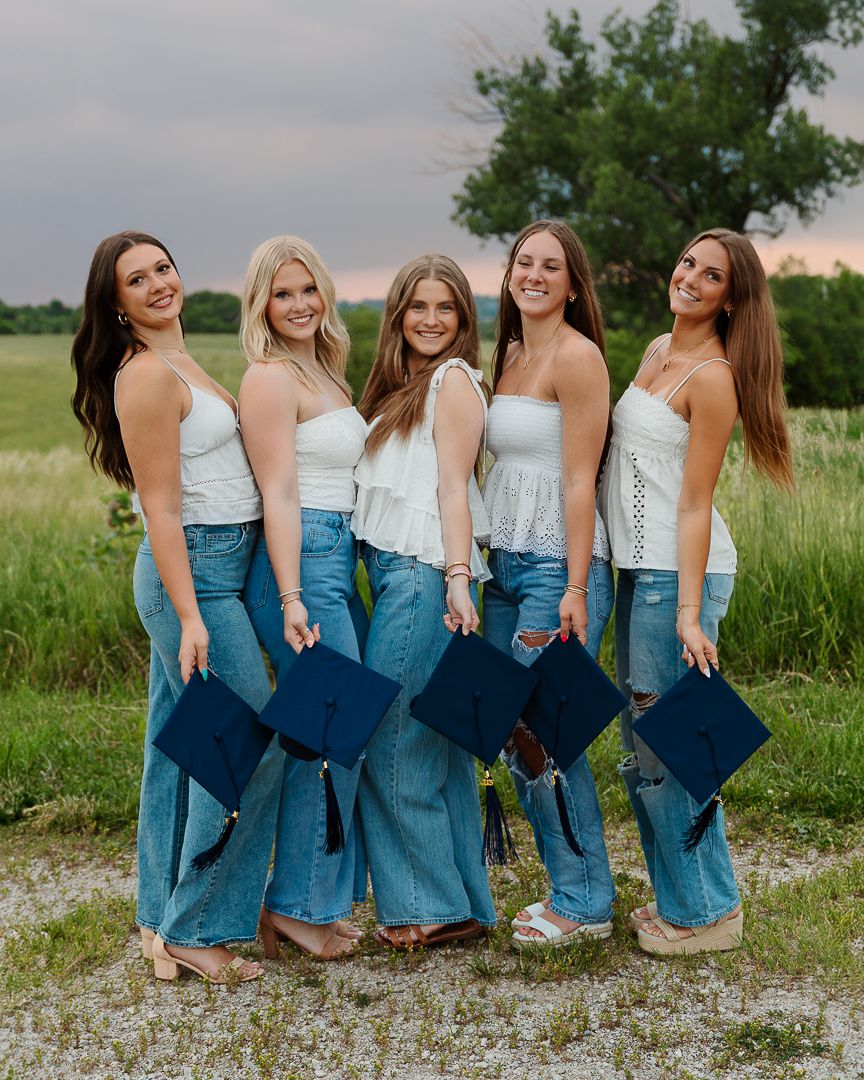 A group of friends in matching white tops and denim skirts pose together on a gravel path surrounded by green grass.