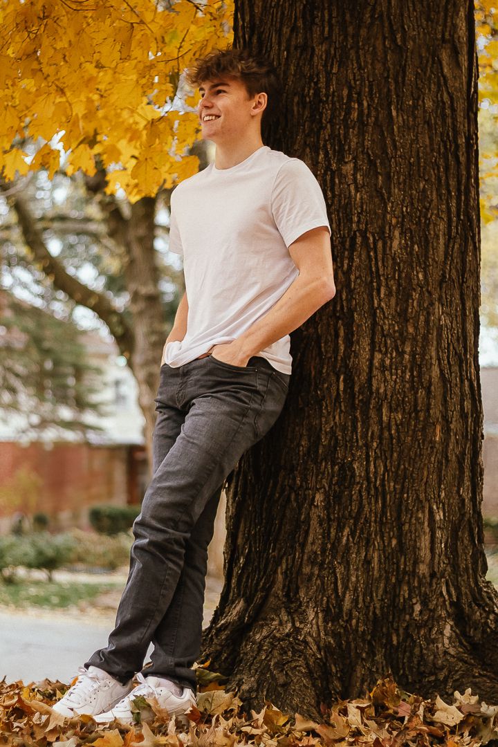 A person in a white t-shirt and dark pants sits against a large tree trunk surrounded by autumn leaves.