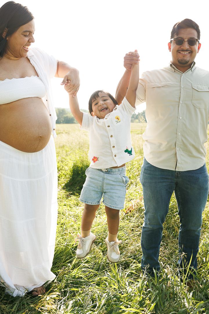 Family wearing white and denim outfits stands together in a sunlit grassy meadow during a casual outdoor photo session.