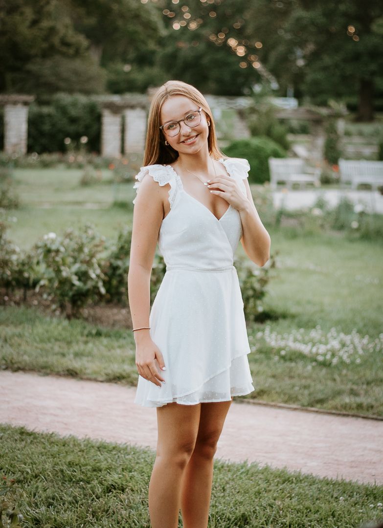 A person in a white summer dress stands on a brick path surrounded by lush green gardens.