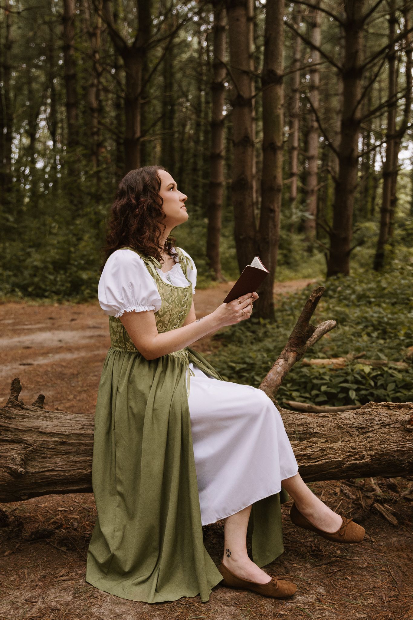 Woman in green medieval dress sitting on a log reading a book in a forest.