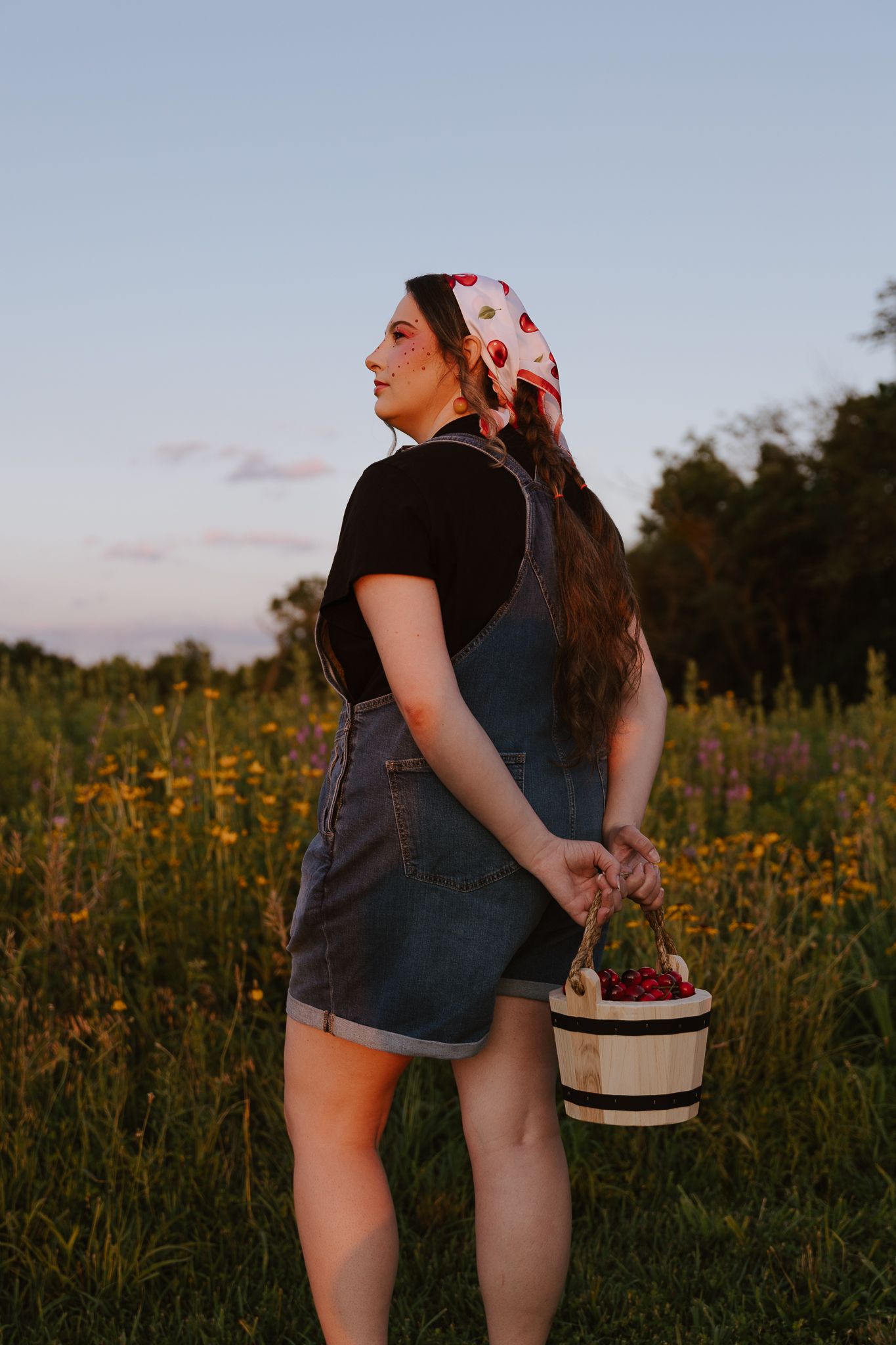 Young woman in denim overalls holds cherry basket while standing in a wildflower field at sunset.
