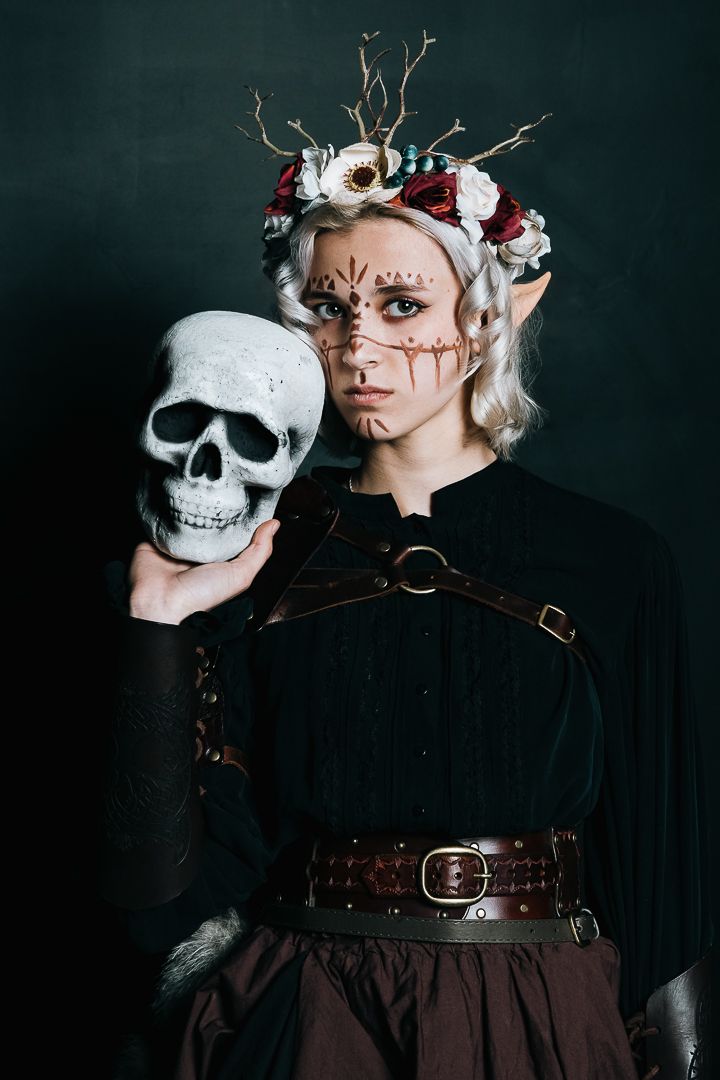 A dark artistic portrait featuring an elf-like figure in gothic attire holding a decorative skull prop against black background.