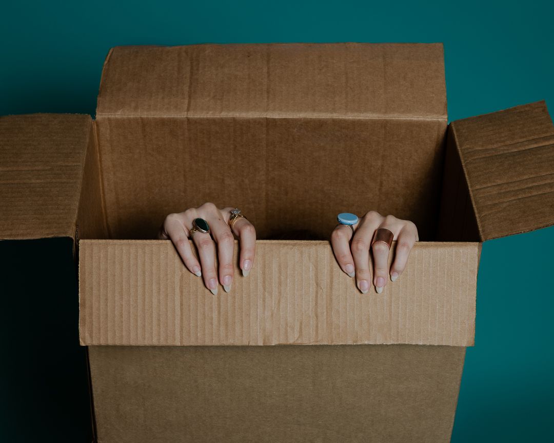 Painted fingernails gripping the edge of a cardboard box against dark teal background.