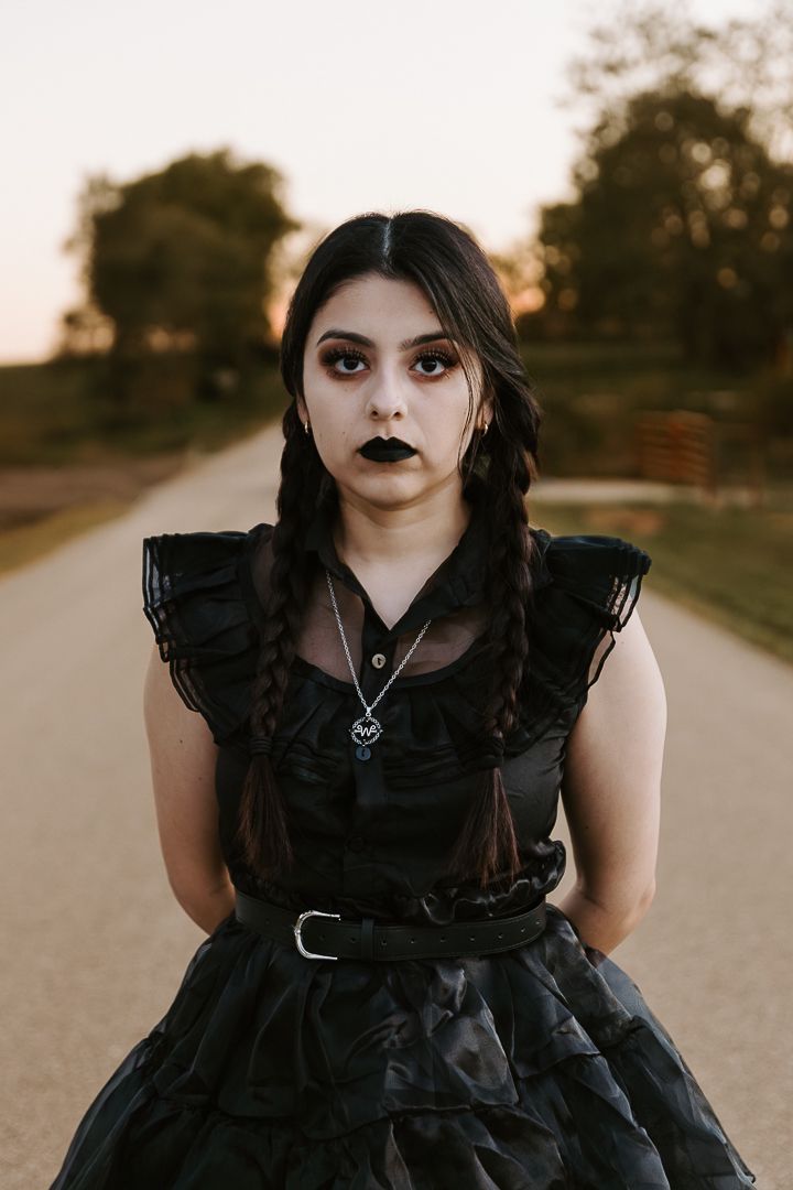 A person in a black gothic dress and dark makeup poses dramatically on a dirt path at sunset.