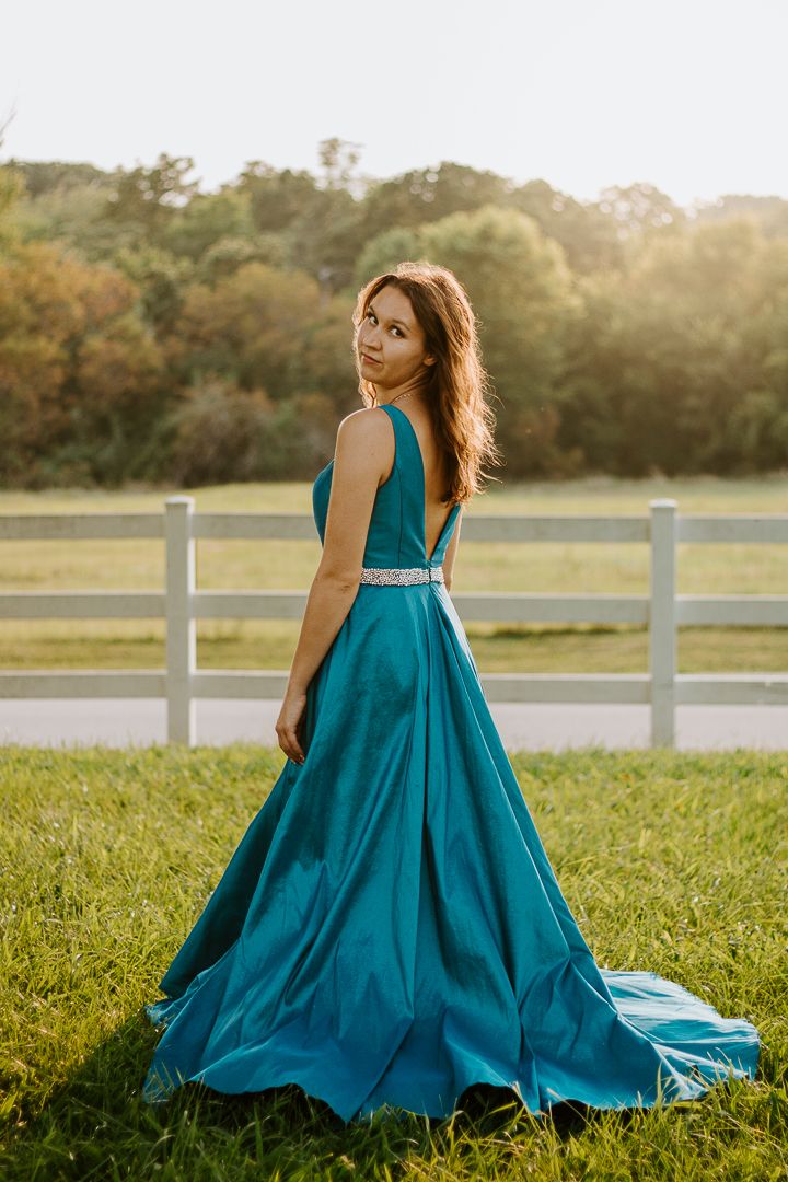 Young woman in teal ball gown with jeweled belt poses gracefully outdoors near white fence at golden hour.