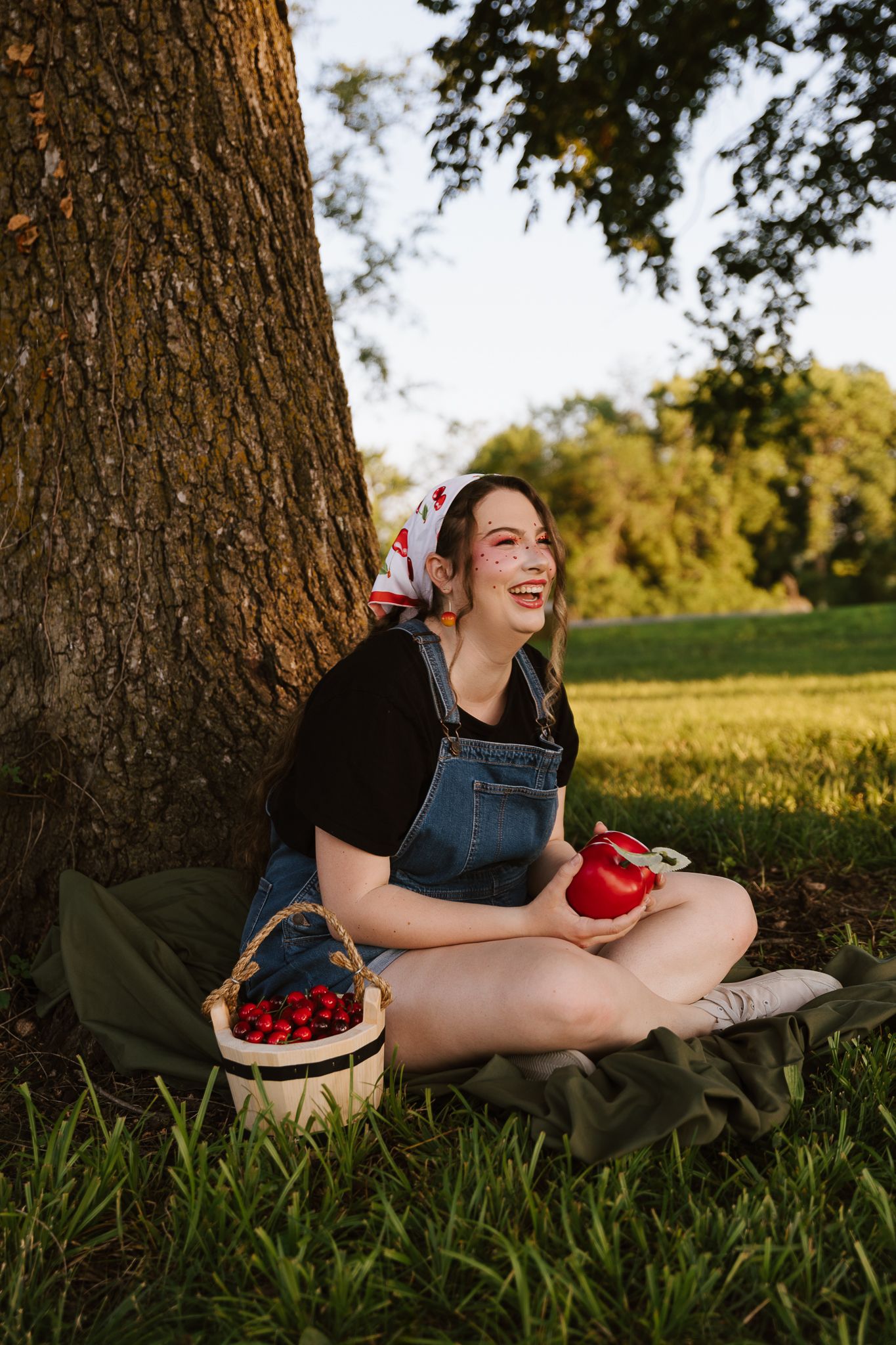 Smiling woman sits under a tree holding a red apple beside a cherry basket in golden sunlight.