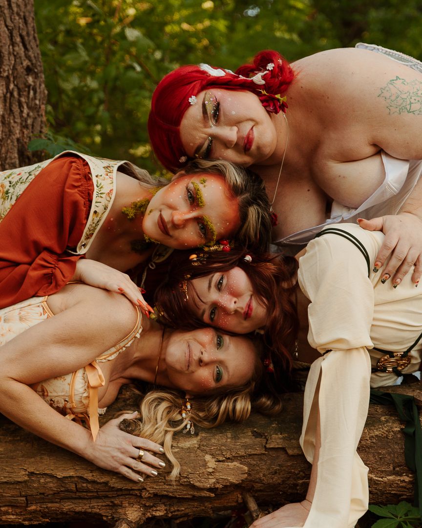 Four women in Renaissance-style costumes pose together on a log in a forest setting.