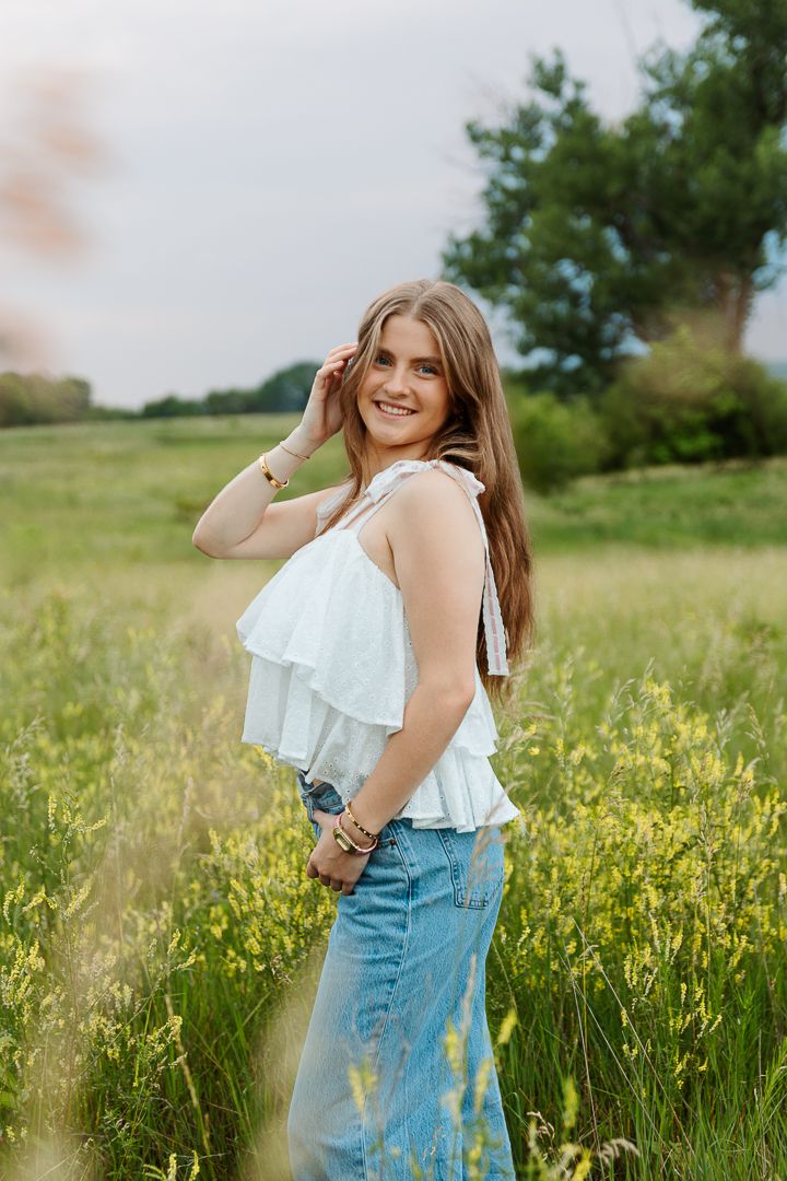 A woman in a white ruffled top and denim skirt poses in a field of yellow wildflowers during golden hour.