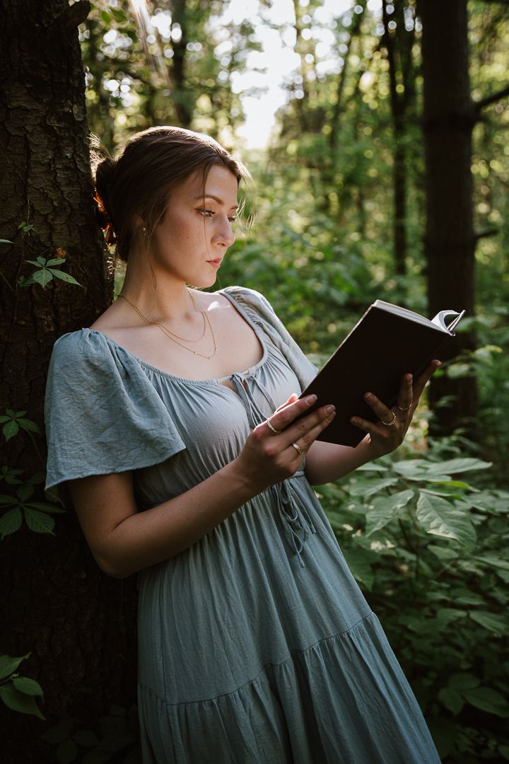 A person in a flowy dress reads a book against a tree trunk in a lush forest setting during golden hour.
