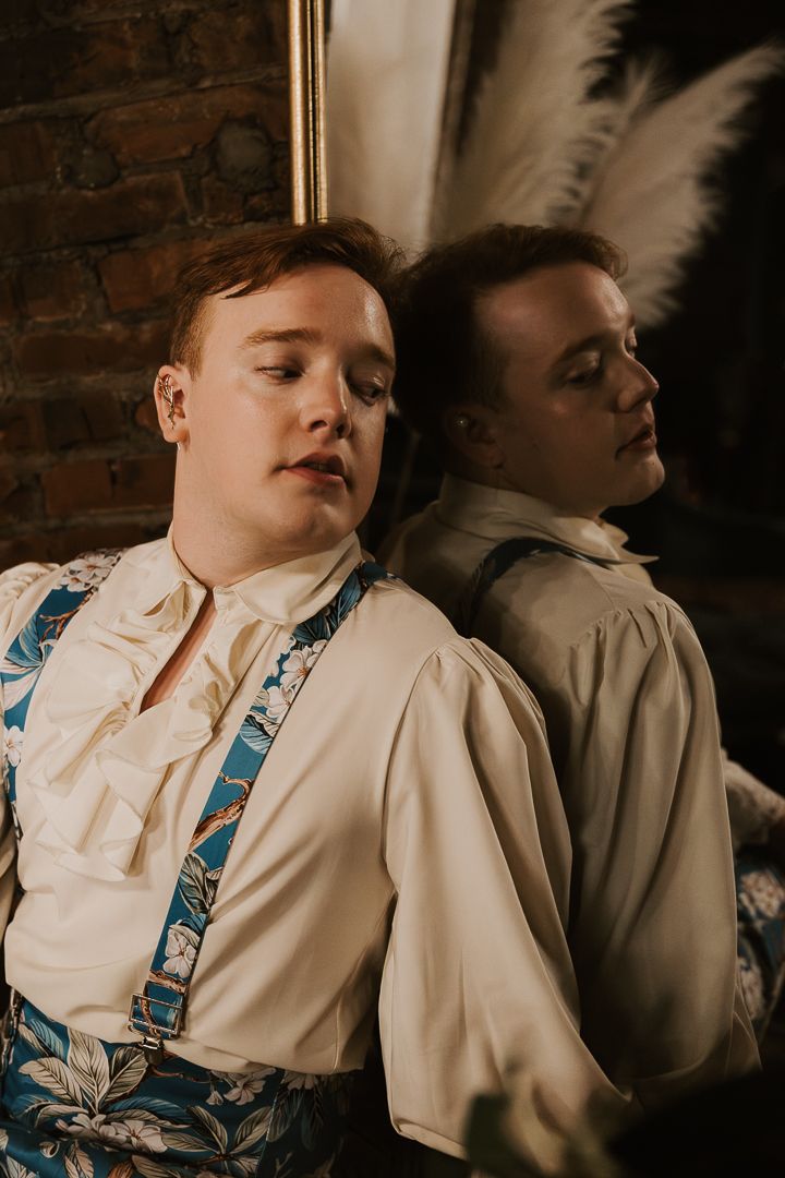 Person in cream colored shirt and patterned vest leans back dramatically against reflective surface in moody lighting.