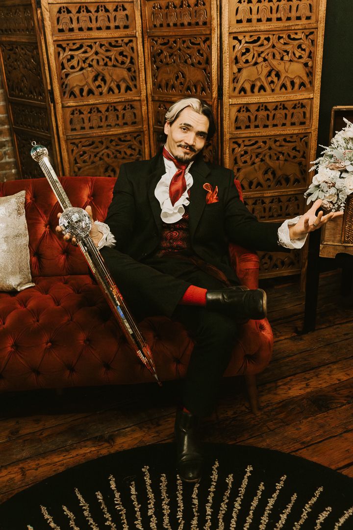 Victorian-style portrait featuring an elegant figure in black suit with red accents seated on a plush red velvet chair.