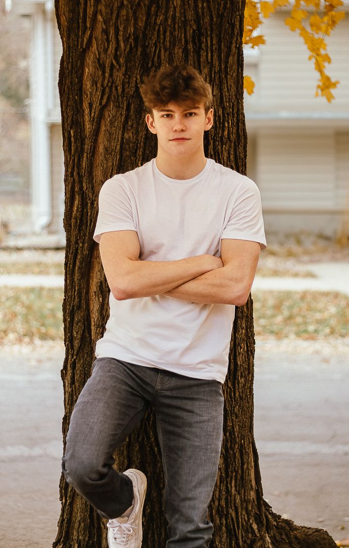 Person leaning against large tree trunk wearing white shirt and dark pants, surrounded by fall leaves.