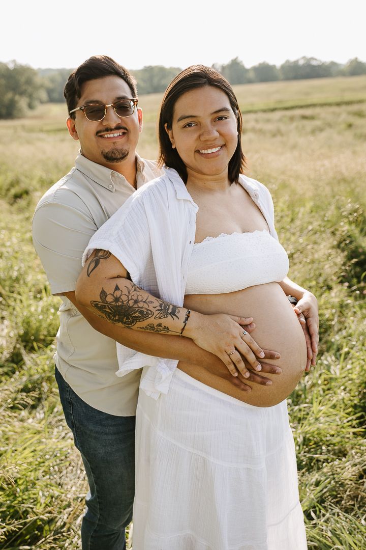 A couple embraces in a sunlit meadow during a maternity photoshoot wearing white and casual attire.