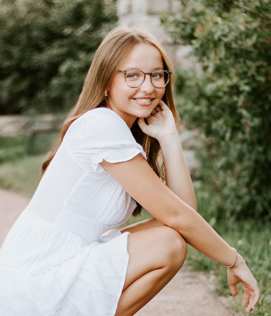 Side view of someone in a white summer dress and glasses posing outdoors near greenery on a sunny day.