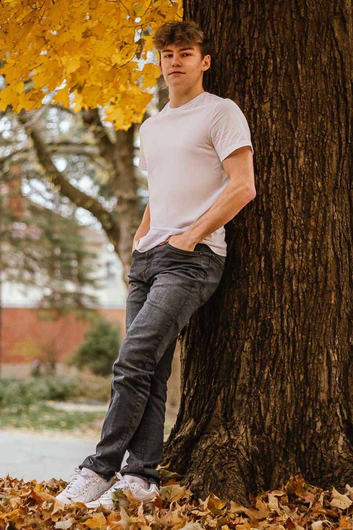 A person in a white t-shirt and dark pants leans against a tree trunk surrounded by golden autumn leaves.
