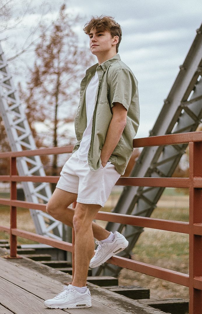 Person wearing olive vest and white shorts sits casually on wooden railing of rustic bridge structure.