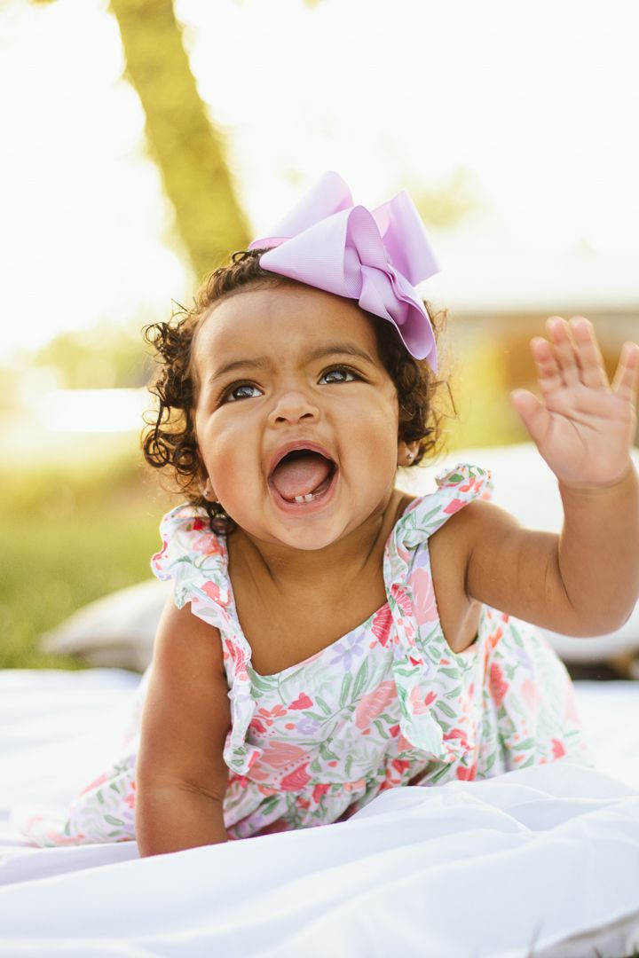 Baby wearing floral dress and purple bow smiles brightly while playing on white blanket outdoors.