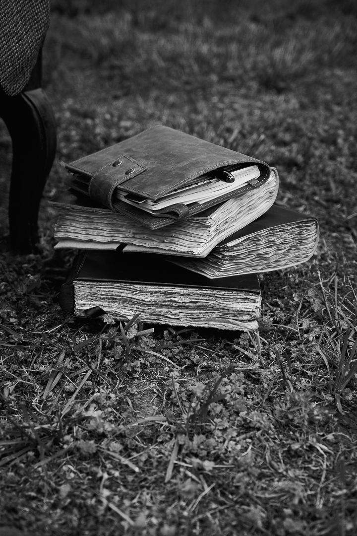 Black and white photo of a stack of old, worn books with a leather journal on top, resting on grass outdoors.