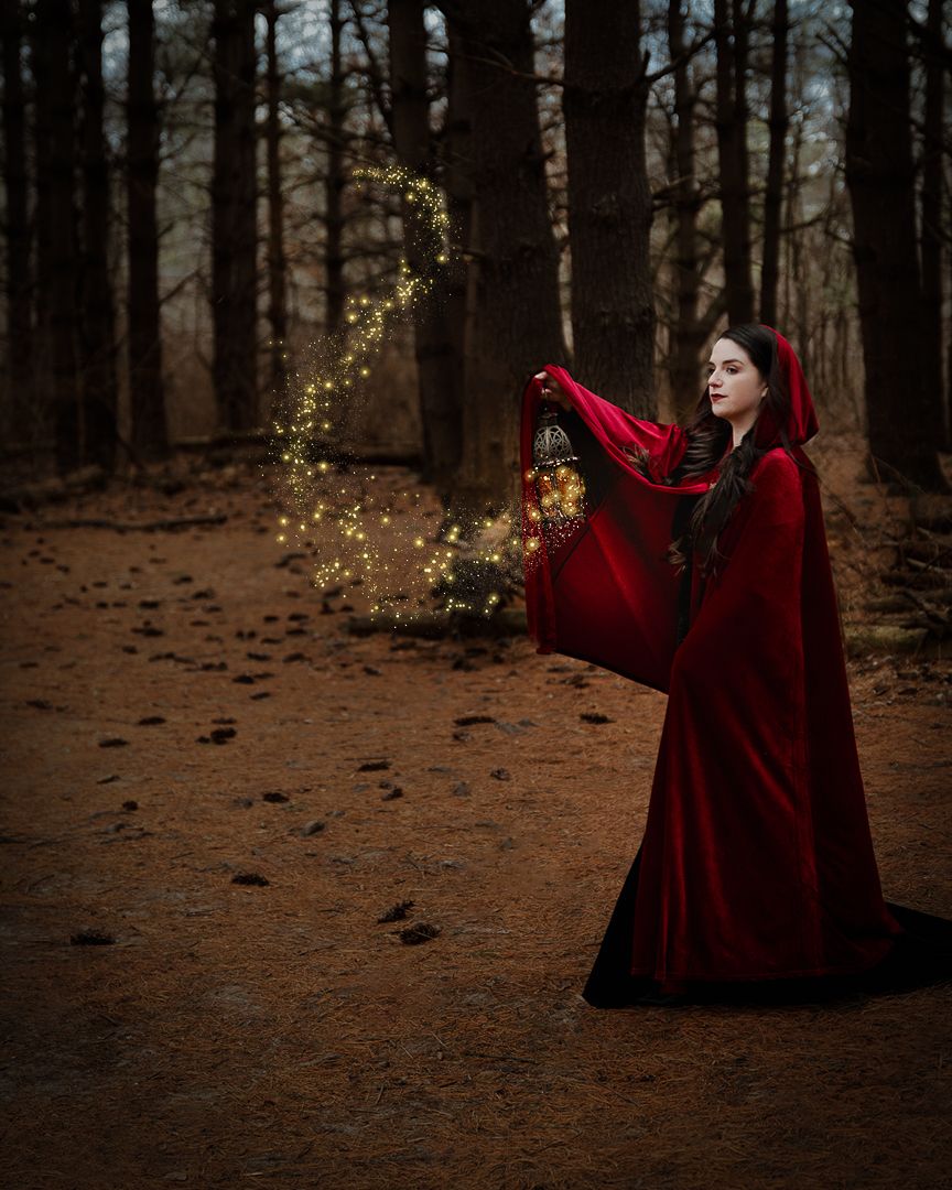 Woman in red hooded cloak holds glowing lantern with magical sparks in a dark forest.
