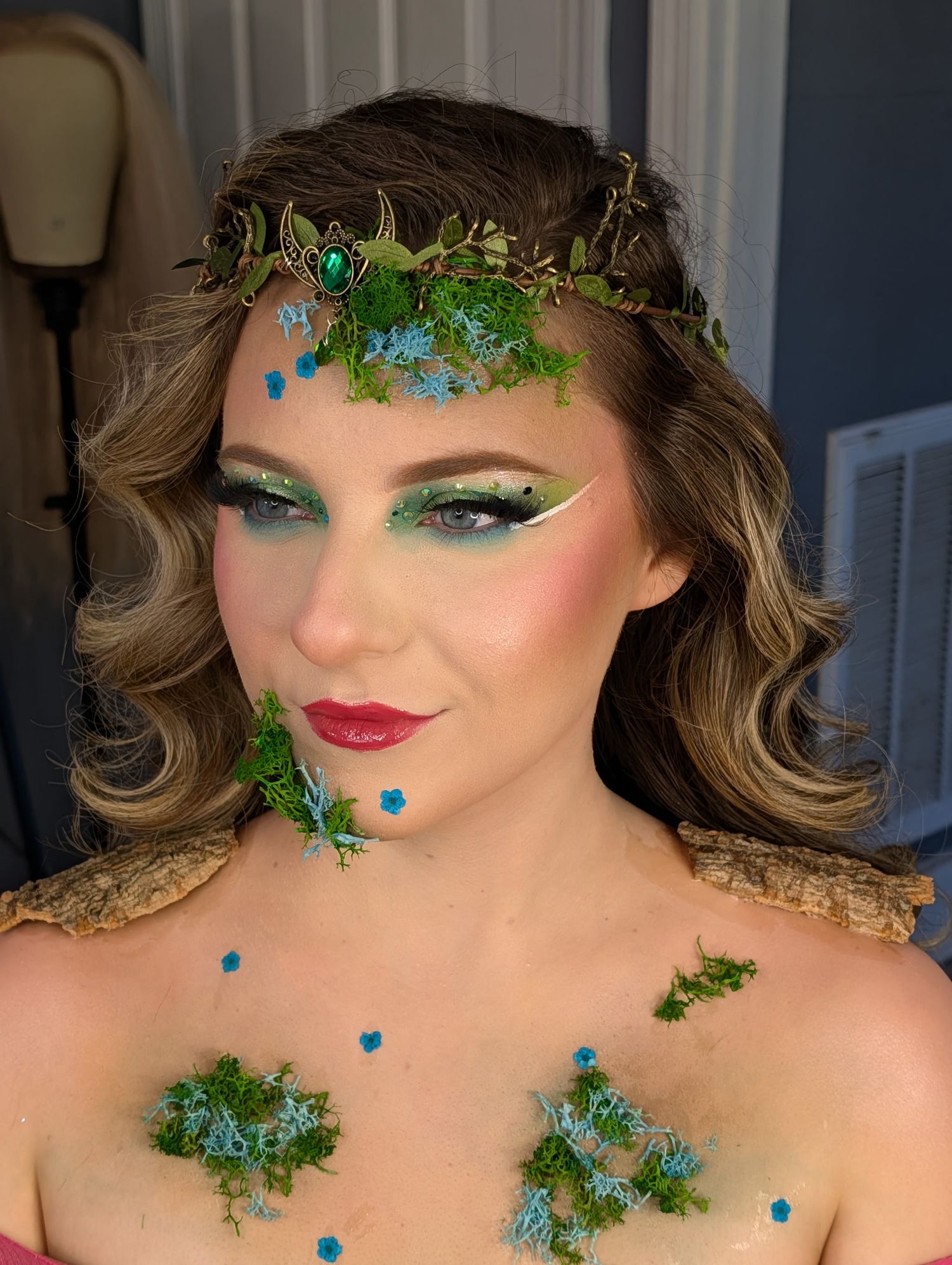 Woman with fantasy forest fairy makeup, green eyeshadow, floral crown, moss decorations, and blue flower accents on face and chest.