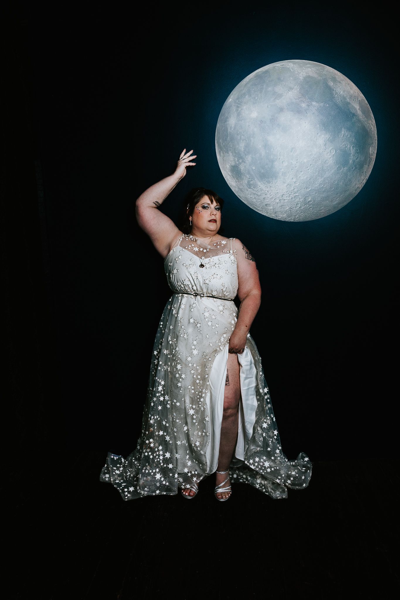 Plus-size woman in sparkly white gown poses dramatically before a glowing full moon on black background.