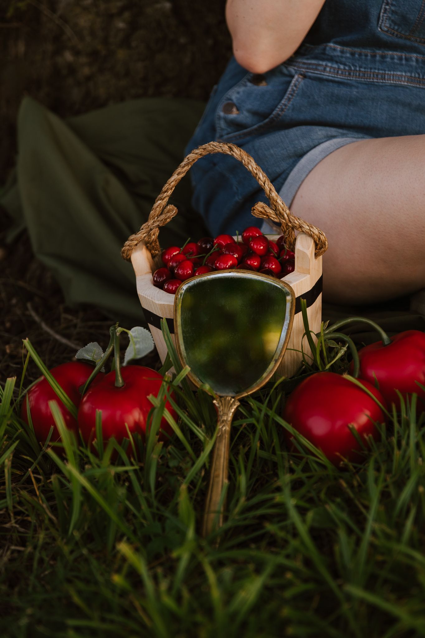 Rustic wooden basket filled with red cherries beside a vintage mirror and apples on green grass.