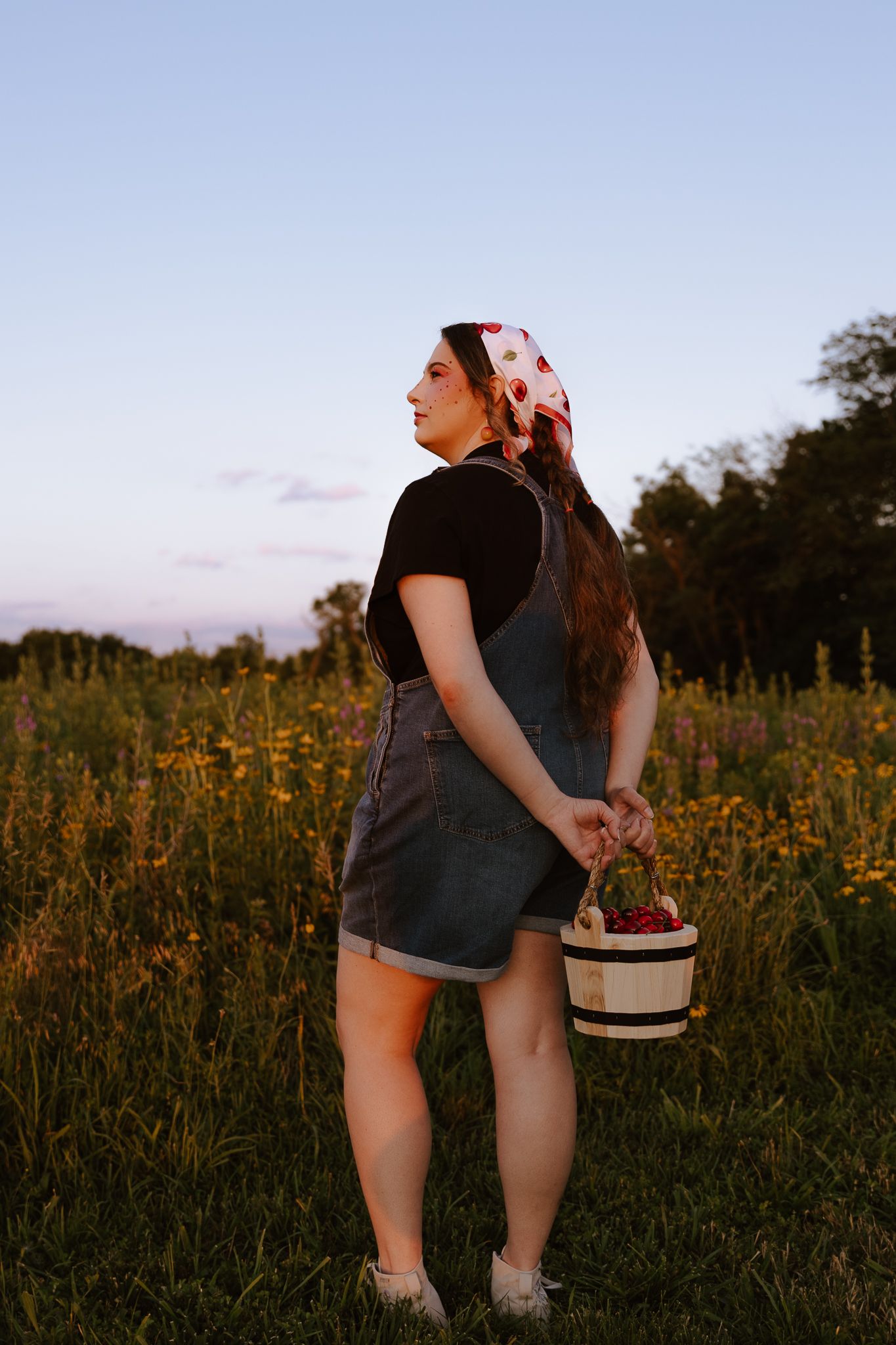 Woman in denim overalls and floral headscarf holding a basket in a wildflower field at dusk.