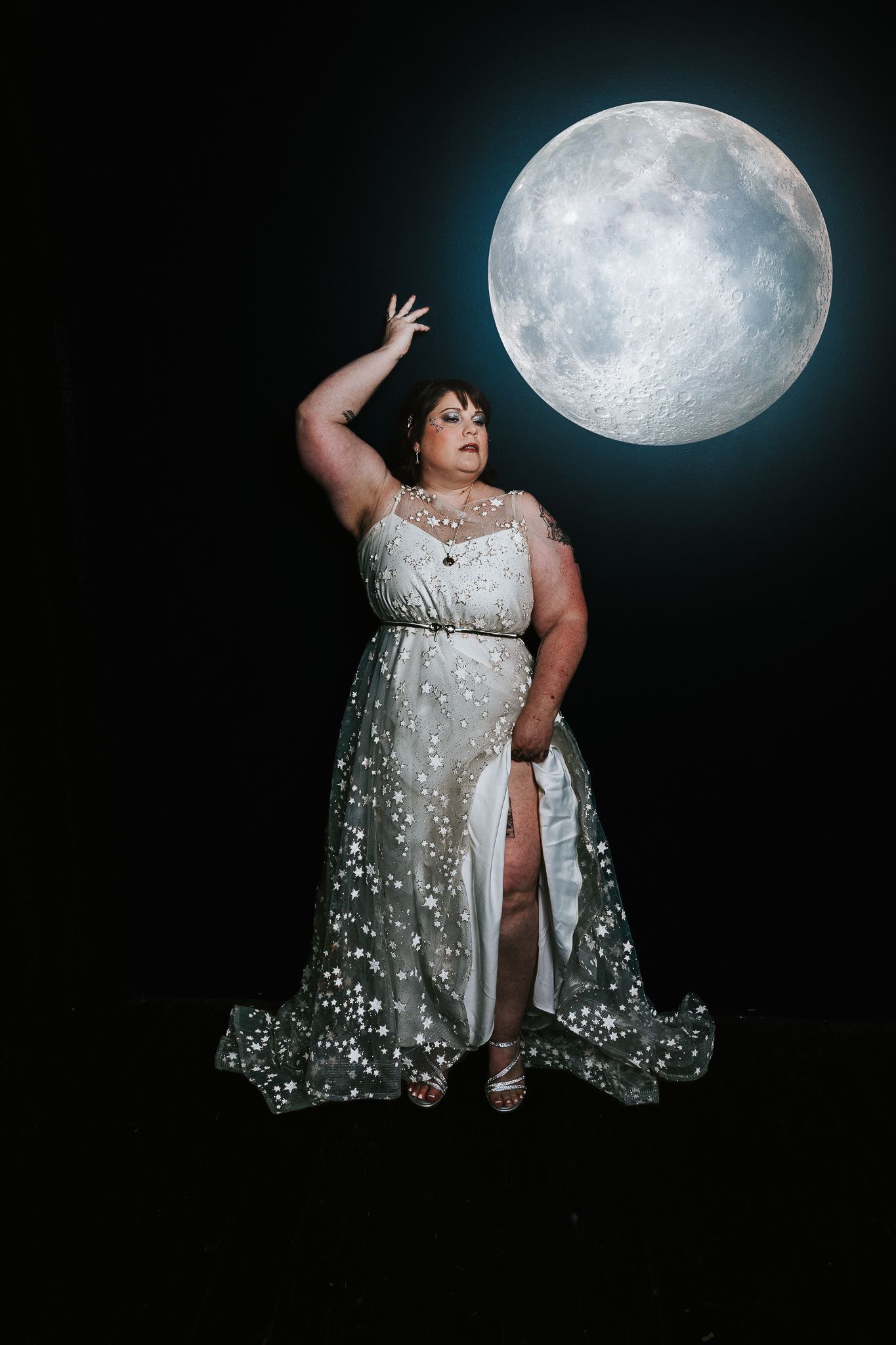 Plus-size woman in silver star-print gown poses dramatically before a glowing full moon on black background.