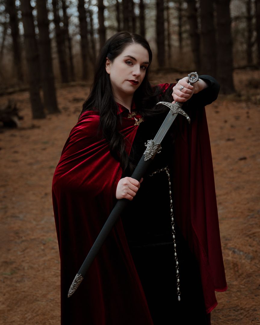 Woman in red velvet cape holding a medieval sword in a forest, fantasy cosplay portrait.