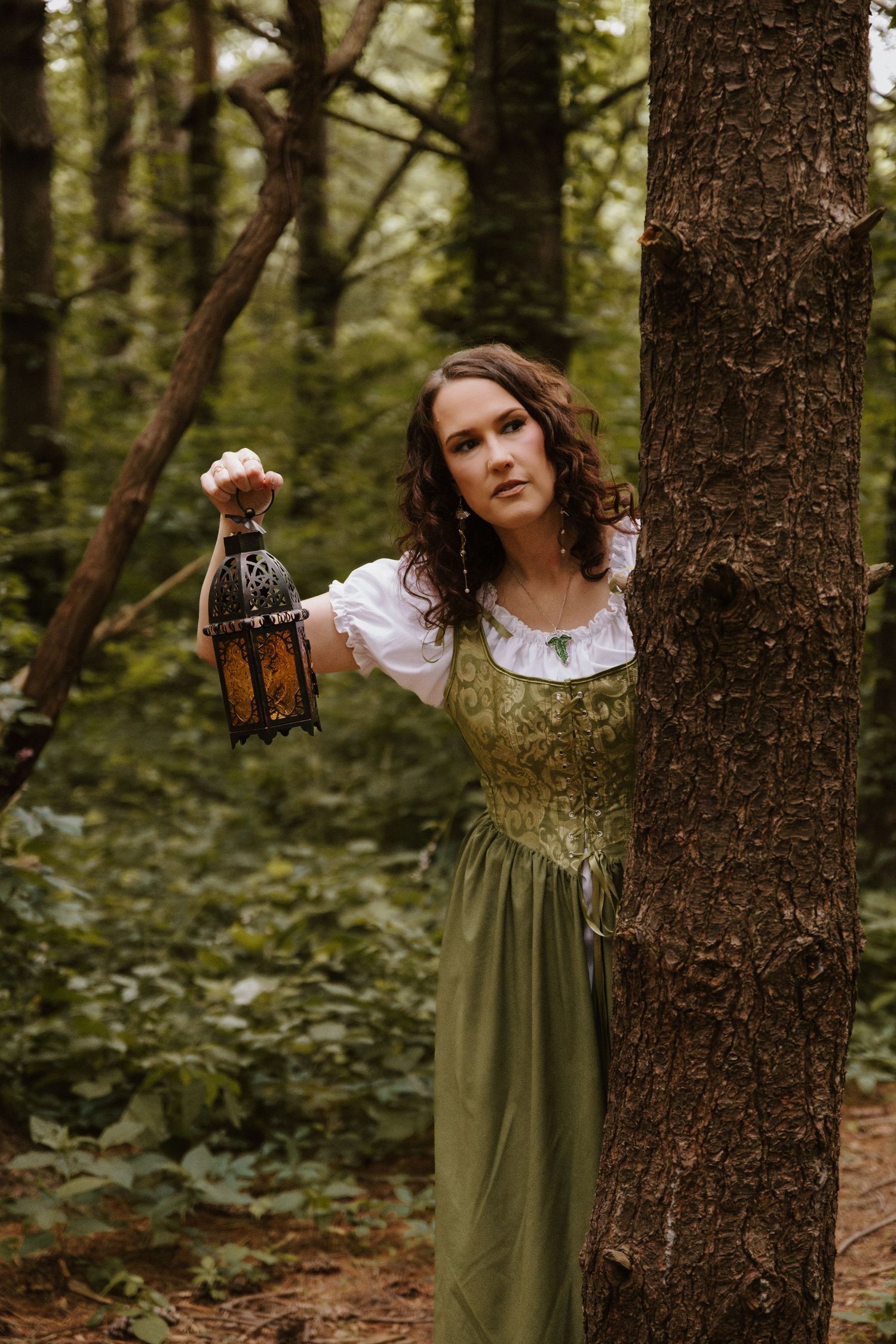 Woman in medieval green dress holds ornate lantern in forest.
