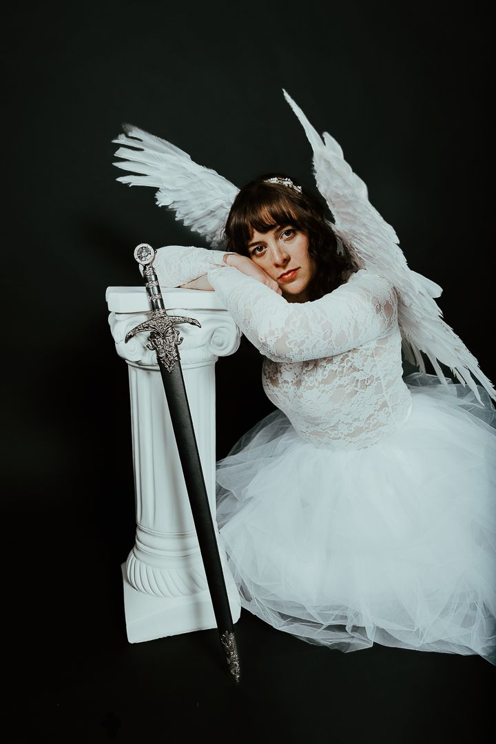 Woman in a white lace gown and feathered angel wings rests her head on a white pillar beside an ornate sword against a black backdrop.