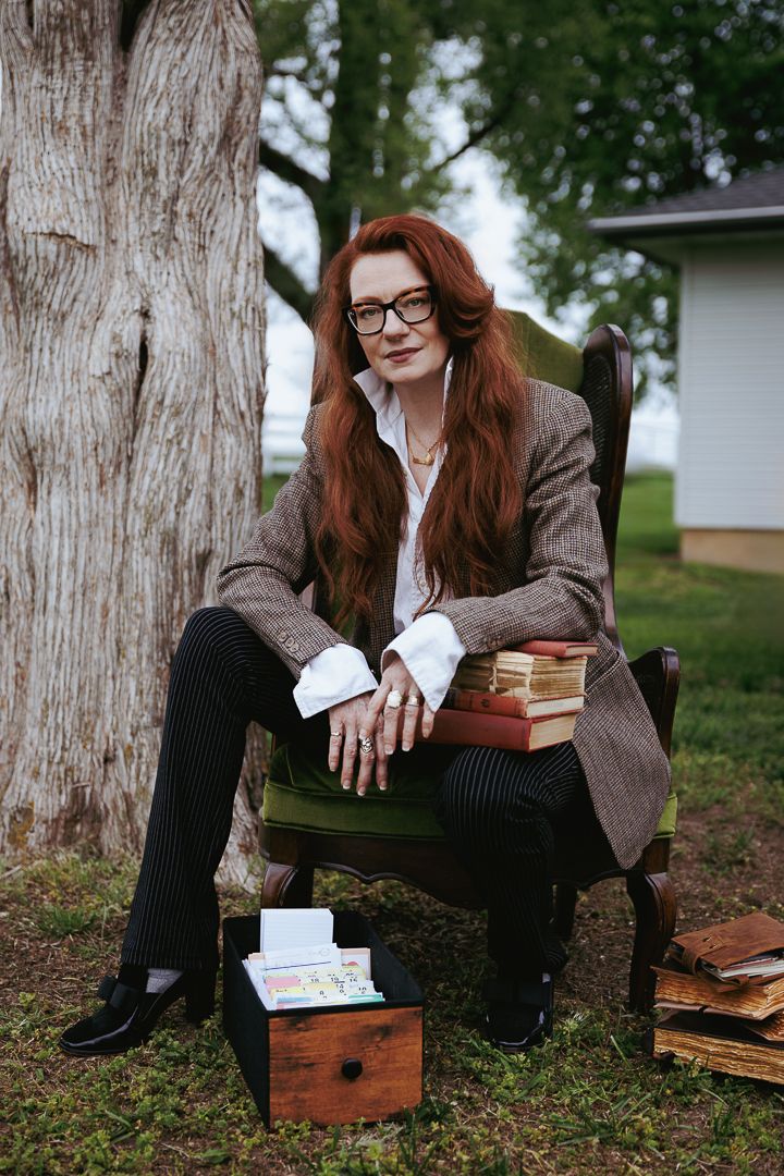 Person in brown coat and glasses sits near tree trunk with books scattered on the ground.