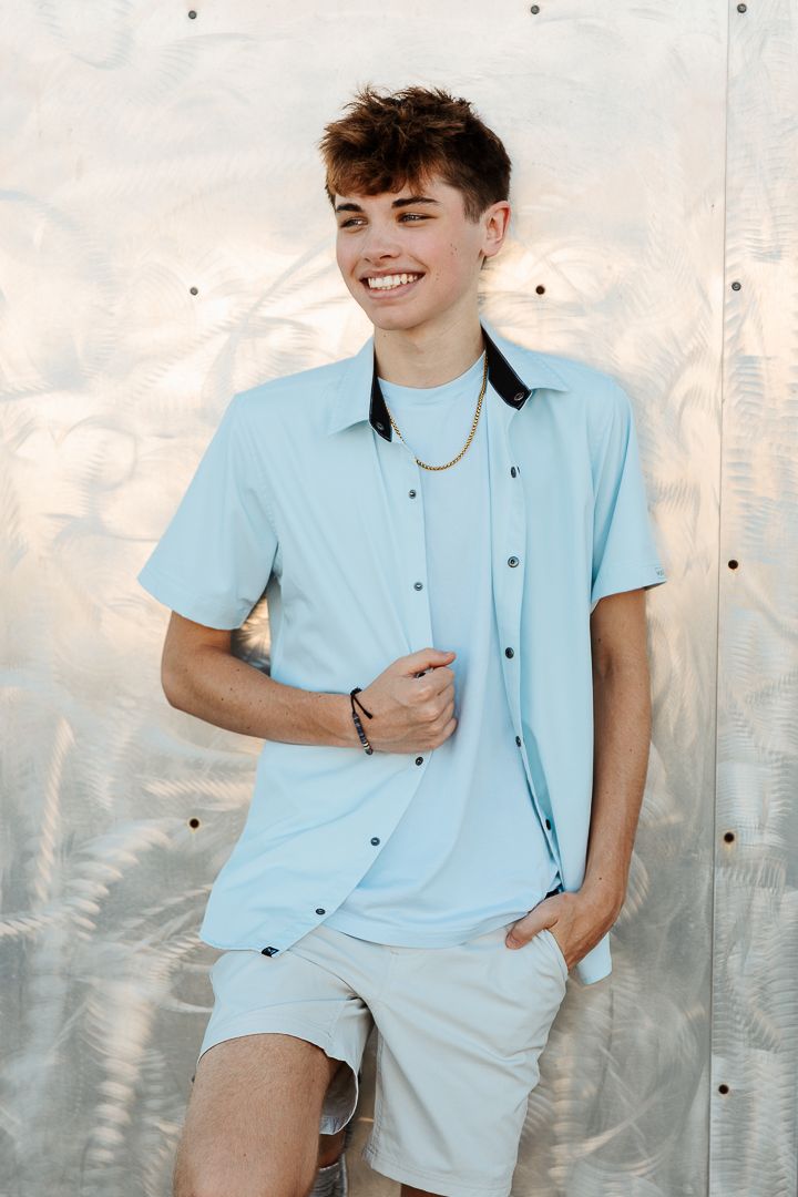 A person in a light blue button-up shirt and white shorts poses against a textured background in bright sunlight.