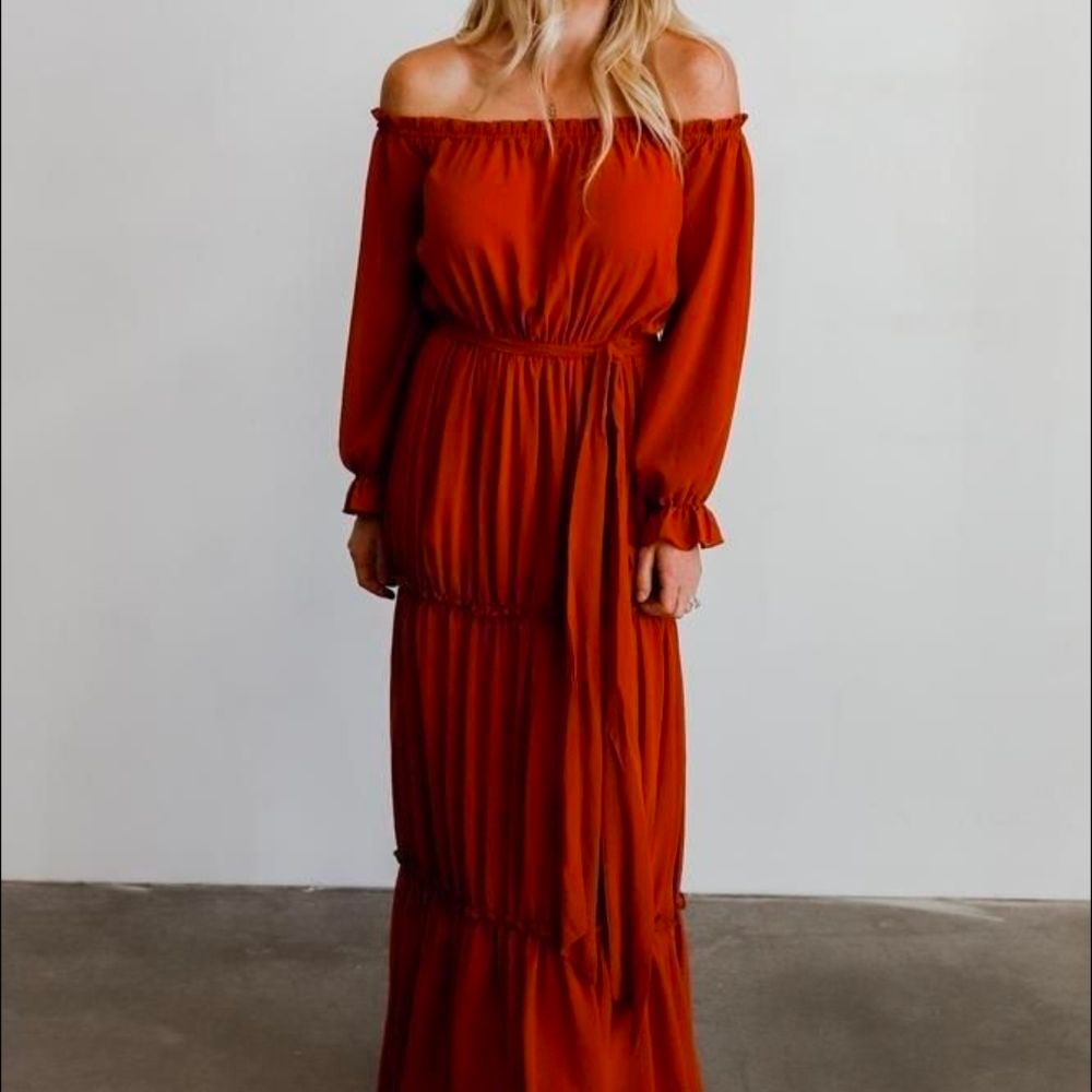 Woman wearing a rust orange off-the-shoulder maxi dress with long ruffled sleeves, tie waist and tiered skirt.