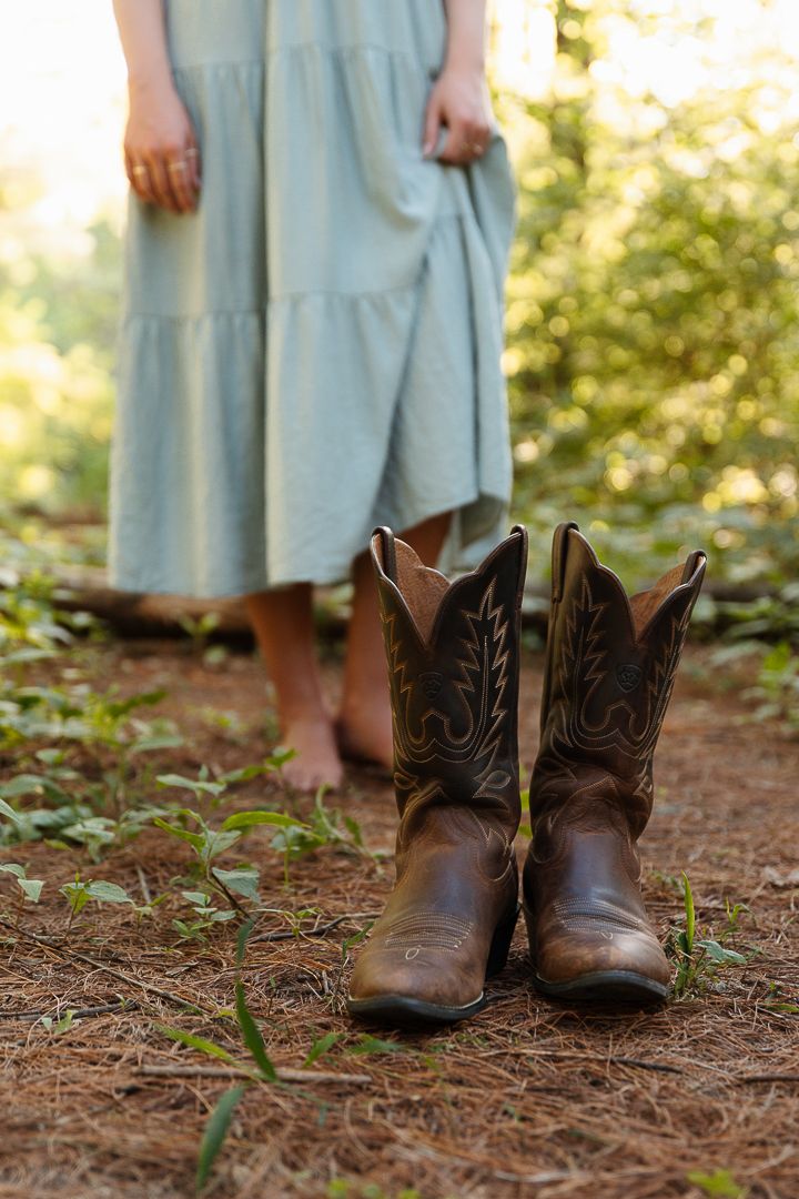 Worn brown cowboy boots on pine needle covered forest floor with sunlight filtering through trees.