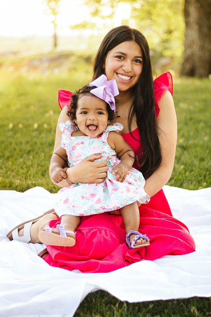 Mother and baby in matching pink and white outfits play and laugh together on a white blanket in a sunny park setting.