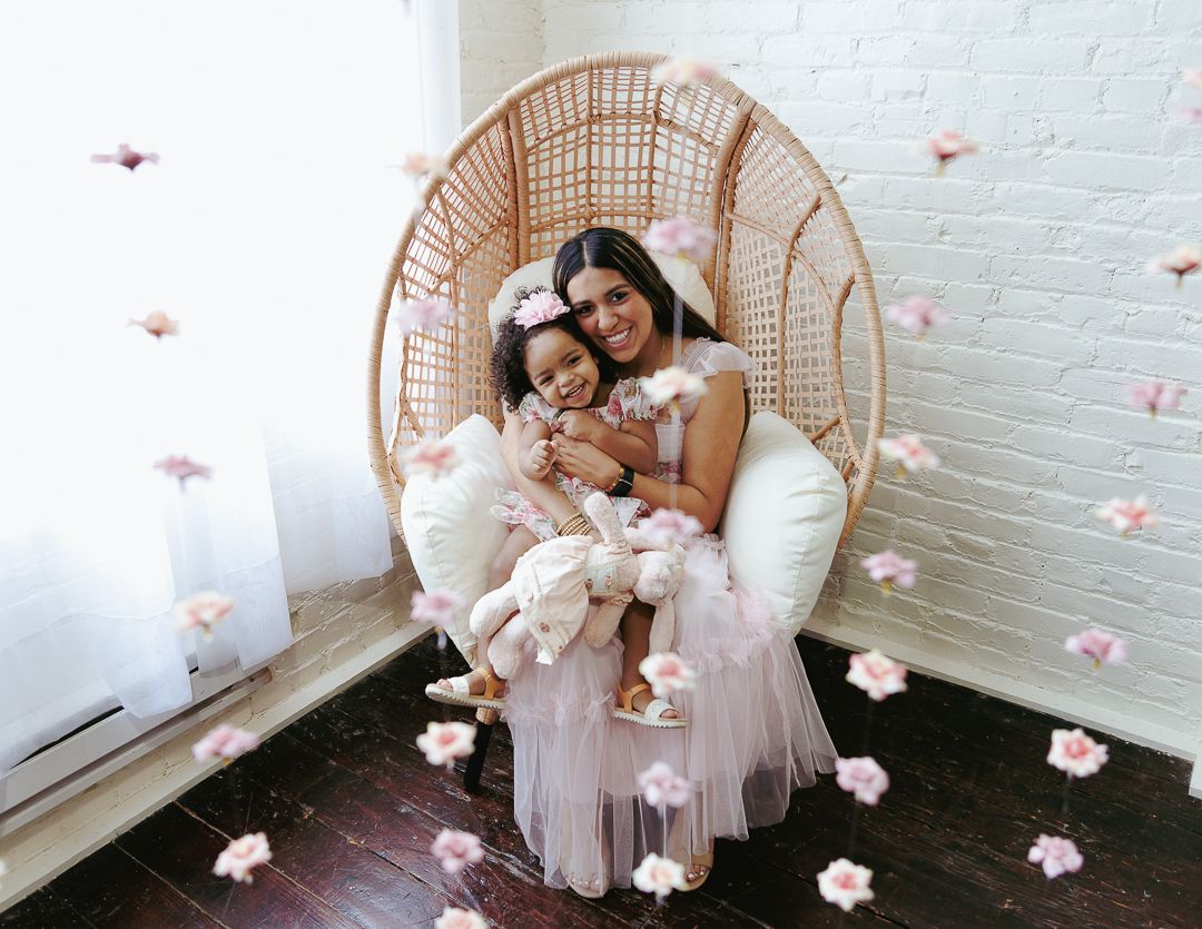 A bride and bridesmaid share a tender moment in a wicker peacock chair surrounded by delicate pink flower petals.
